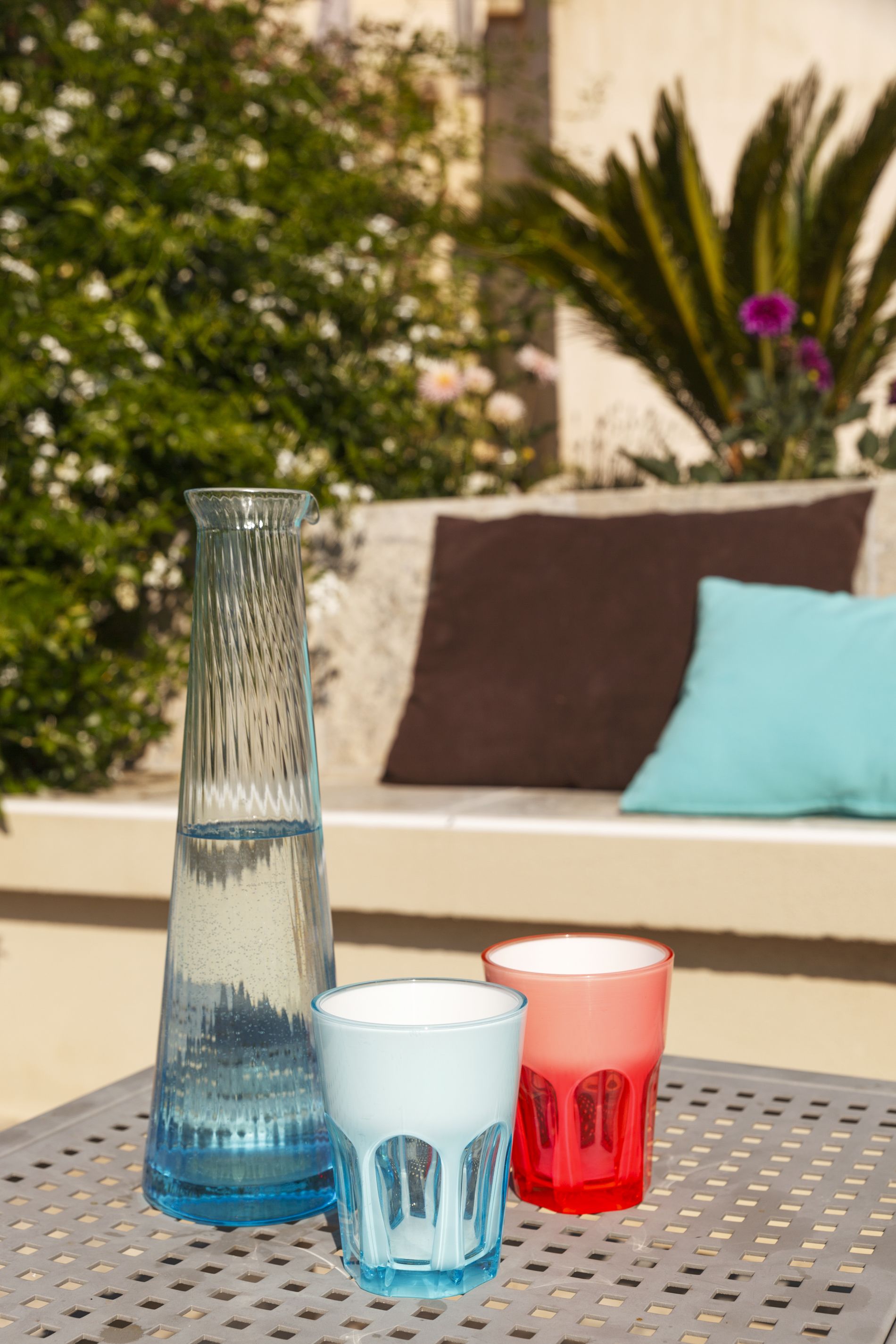 A clear glass vase, a blue glass, and a red glass on a perforated metal table.