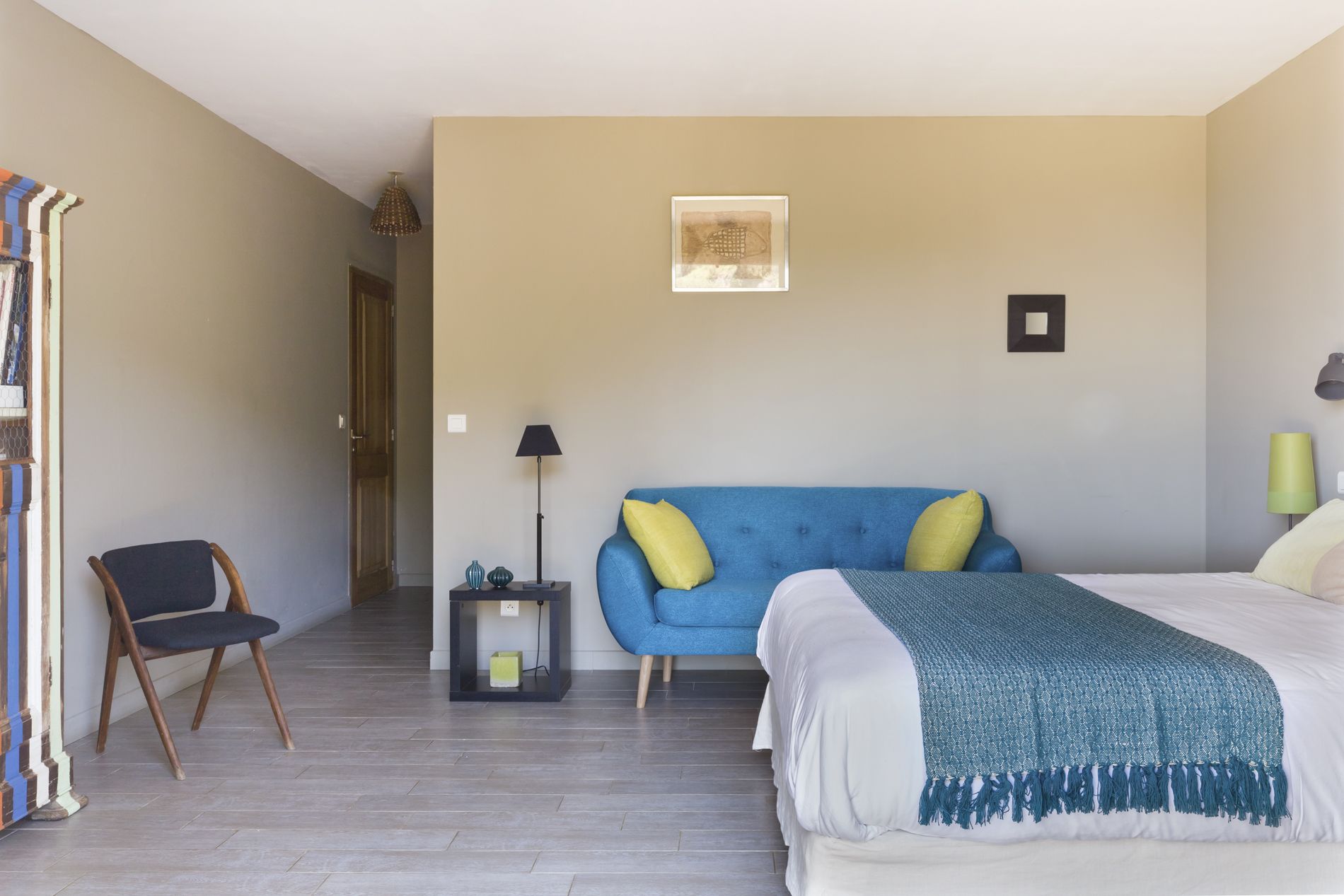 A hotel room with a bed, a blue sofa with yellow pillows, a chair, a lamp, and a framed picture on the wall.