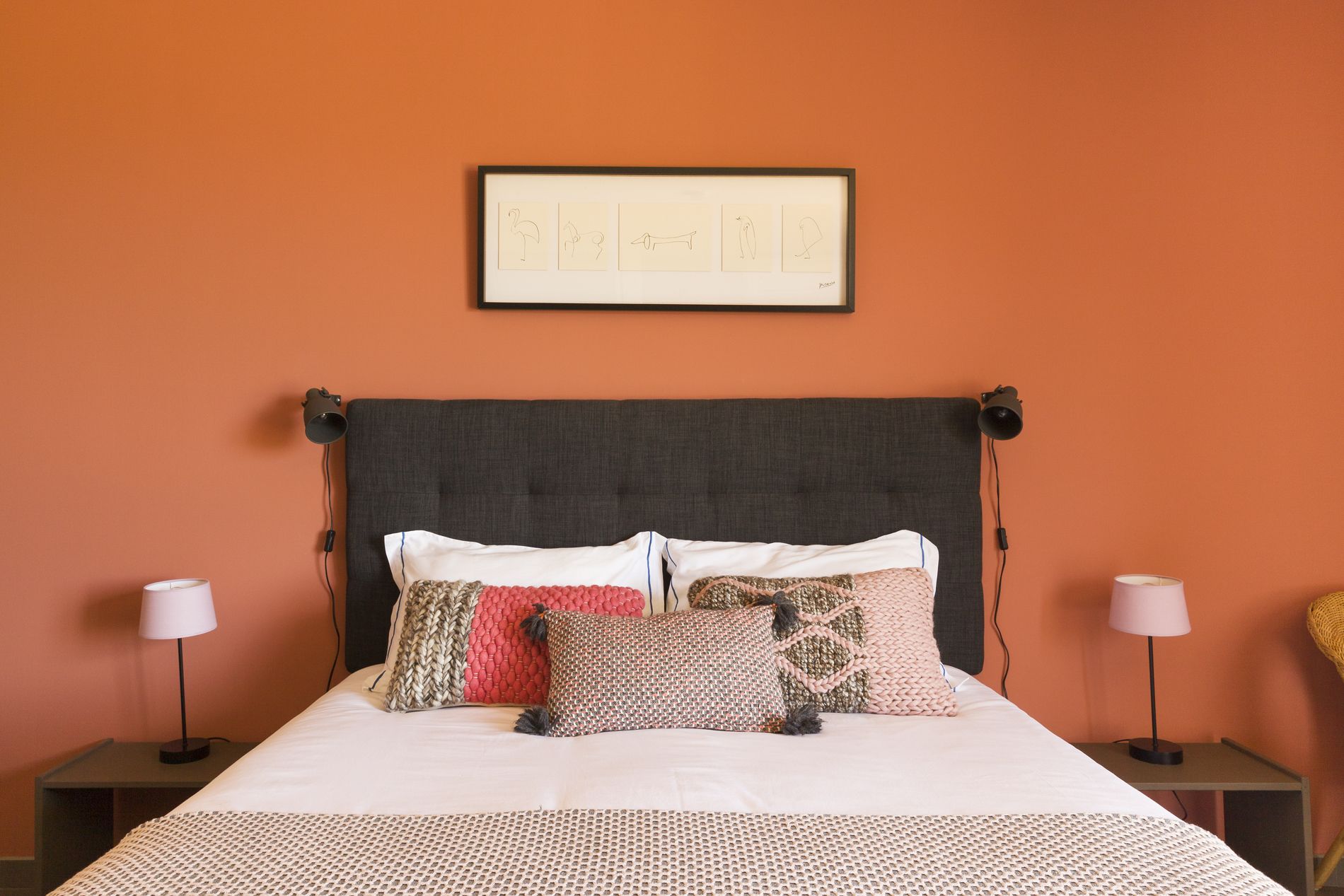 Uma cama com cabeceira cinza, roupa de cama branca e várias almofadas decorativas. Há duas mesas de cabeceira com luminárias, e um quadro emoldurado acima da cama.