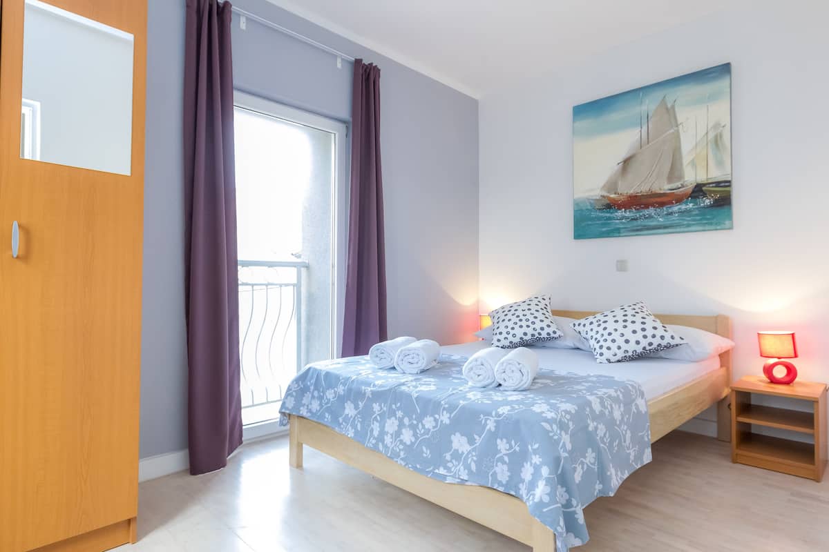 A bedroom with a bed, a painting of a sailboat, a nightstand with a lamp, and a wooden wardrobe.