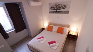 A neatly made bed with white and orange pillows, a window with dark curtains, and a wall-mounted air conditioner. - Apartments in City Center Ventus (Novalja)