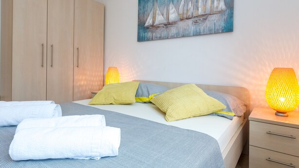 A neatly made bed with pillows and towels, a painting of sailboats, and a nightstand with a lamp. - Maru Apartments (Novalja)