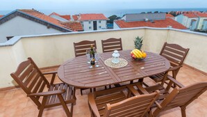 A round wooden table with six chairs, a pineapple, and a bowl of bananas on a balcony with a view of the sea and buildings. - Maru Apartments (Novalja)