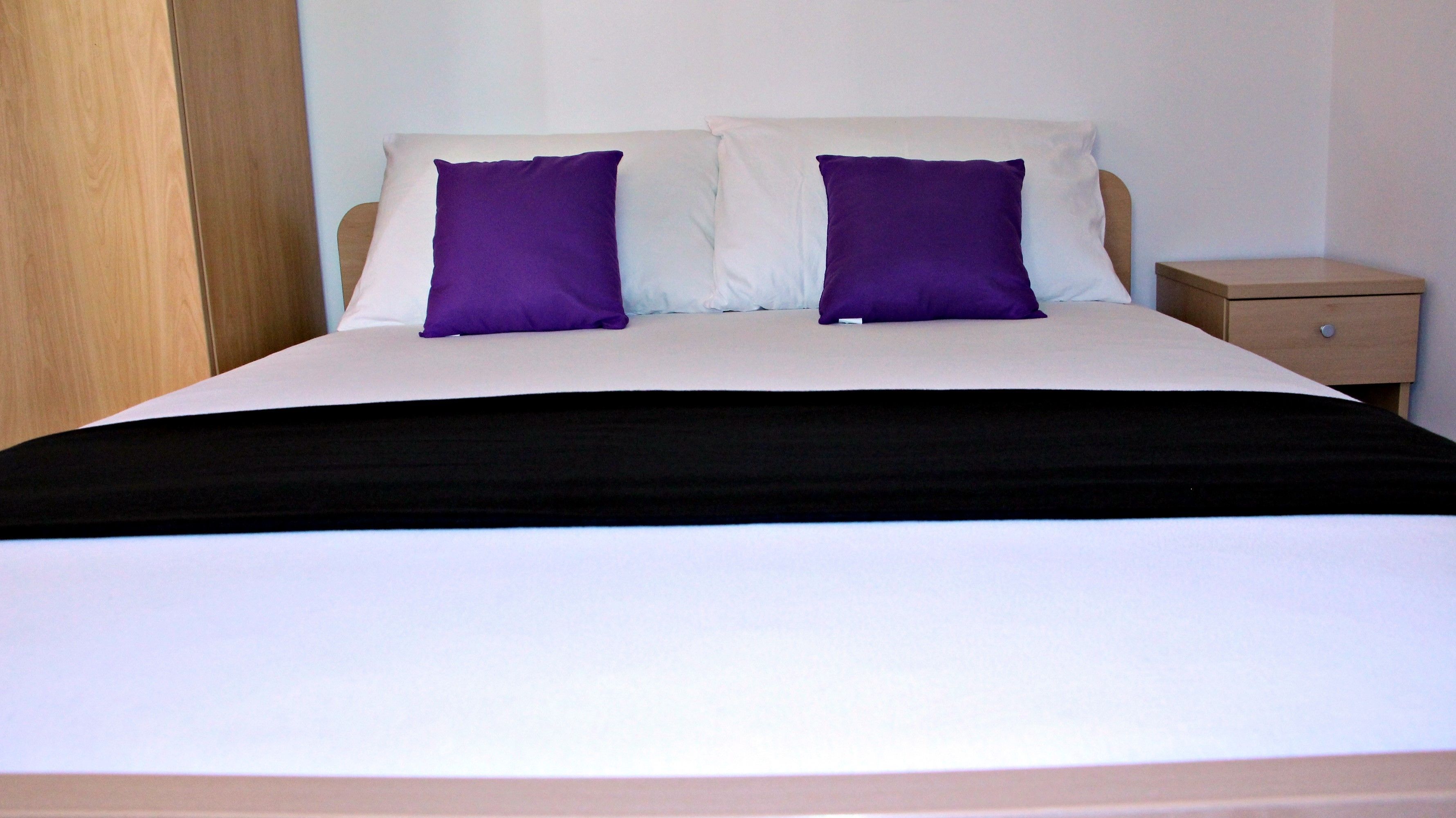 A bed with white and purple bedding, a wooden headboard, and a nightstand with a drawer.