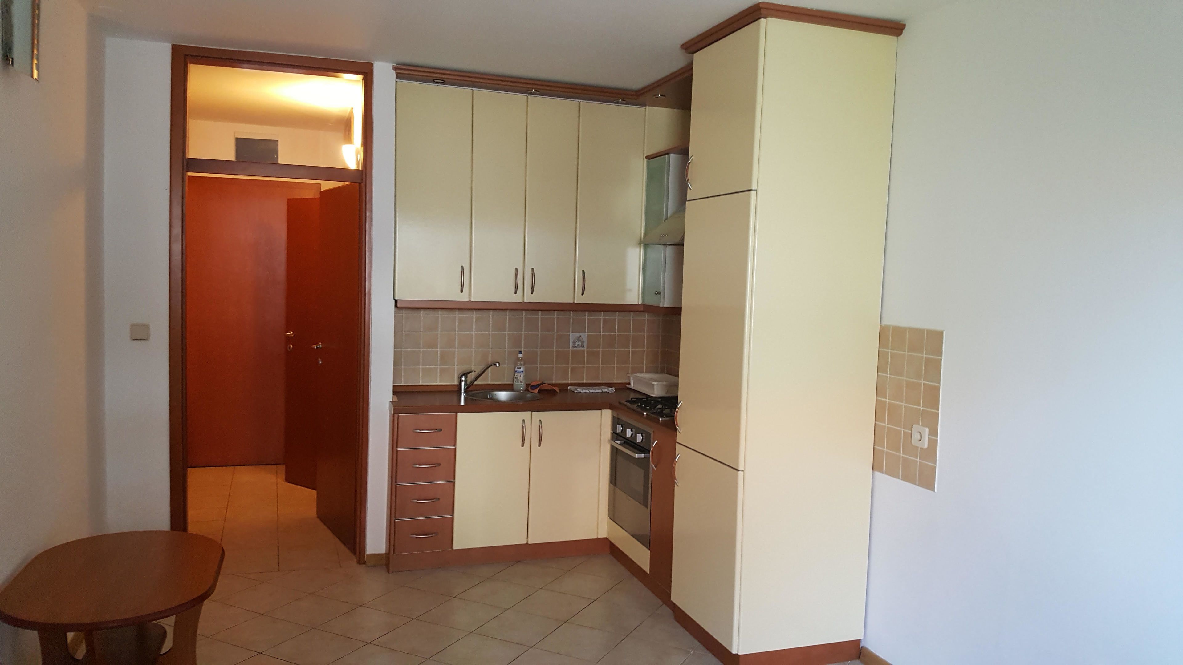 A compact kitchen with a refrigerator, stove, and sink. There is a wooden table and a door leading to another room.