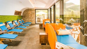 A sunlit indoor relaxation area with blue and orange lounge chairs, a stone wall, and a view of the outdoors. - Guthof Lutz Appartement mit kostenlosen Sommerbergbahnticket Mai bis Oktober (Schattwald)