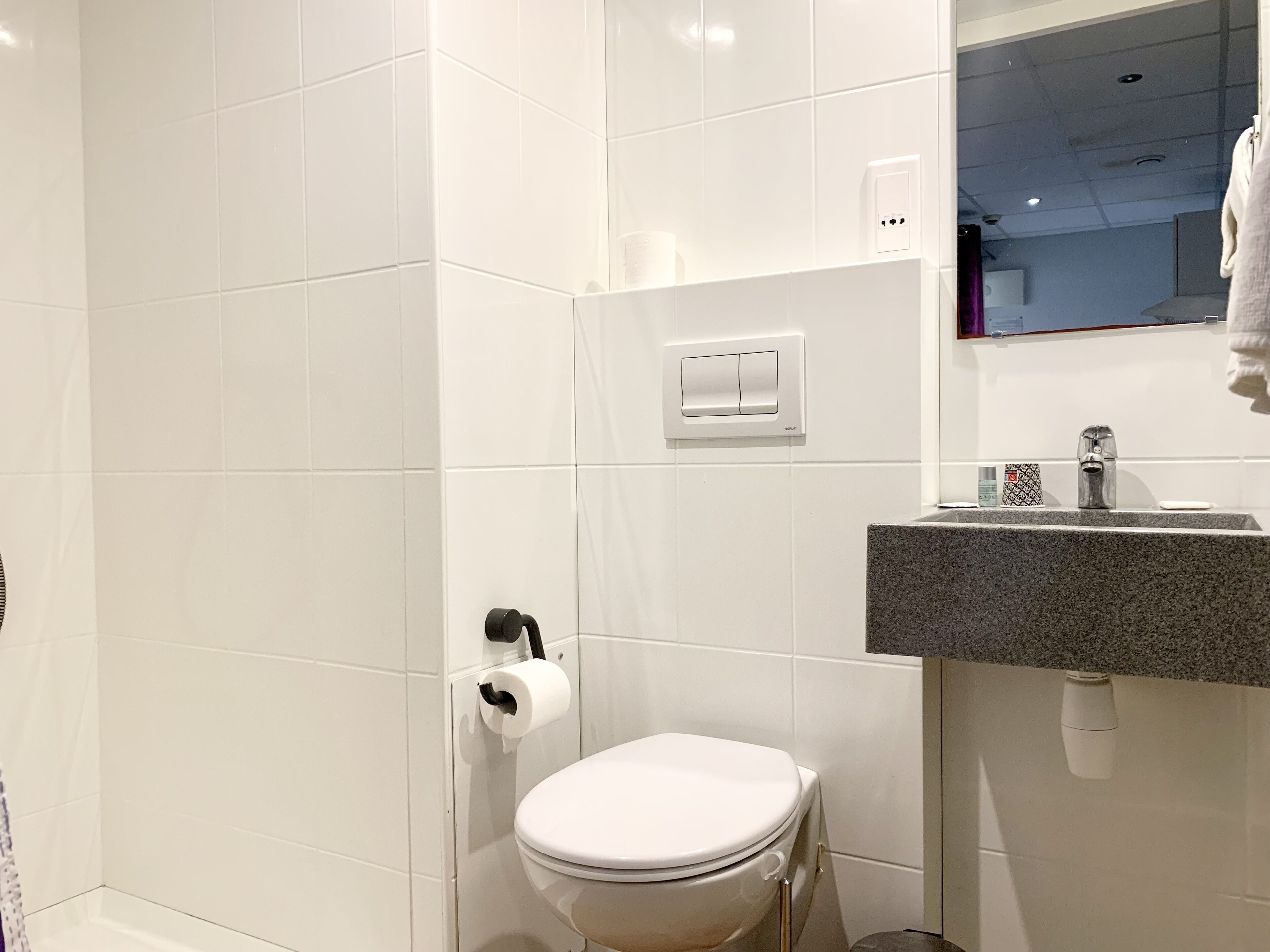 A modern bathroom with white tiles, a toilet, a sink with a granite countertop, and a mirror above the sink.