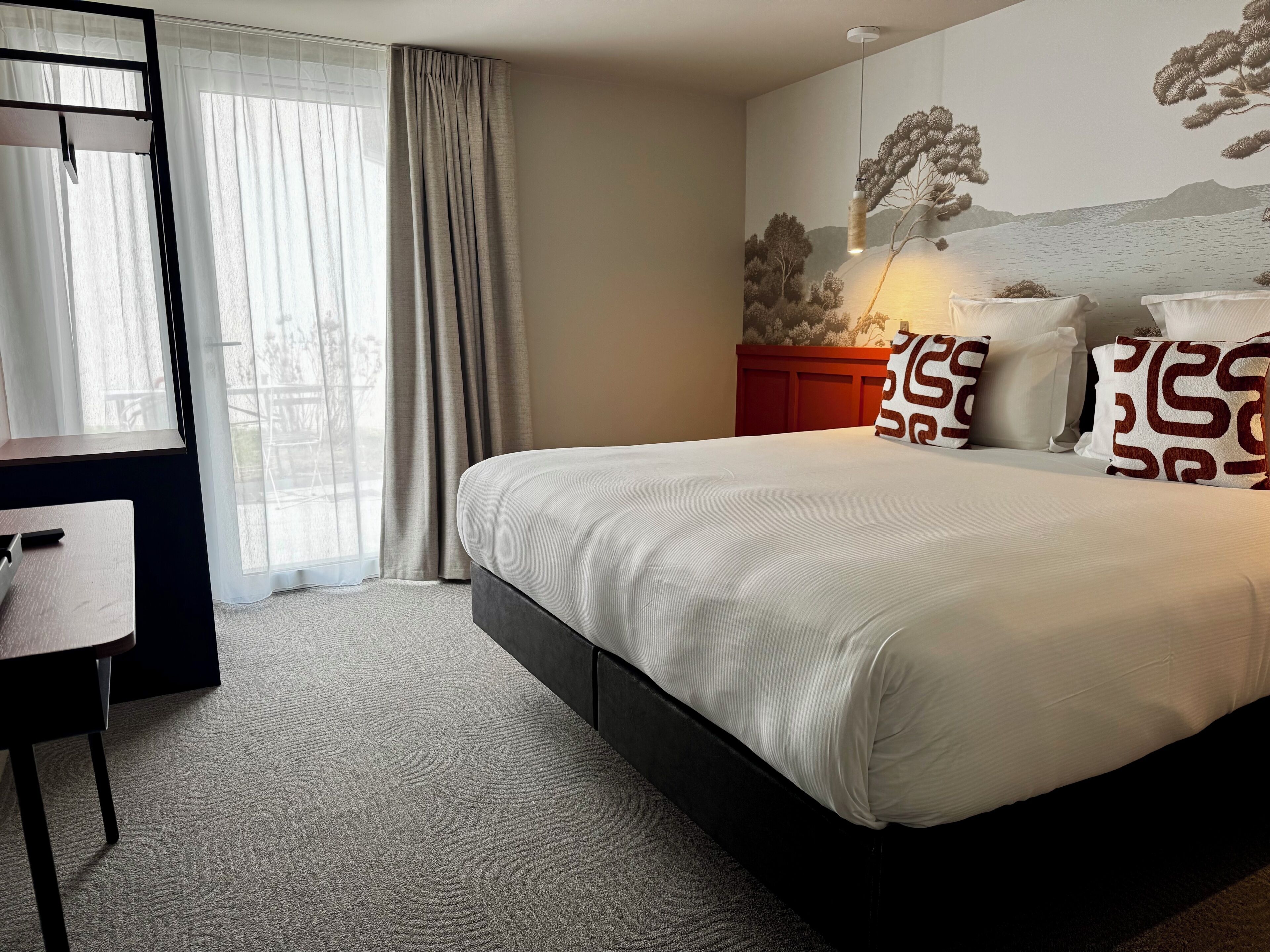 Hotelkamer met een bed, een bureau, een stoel, een rode nachtkastje en een muurschildering.
