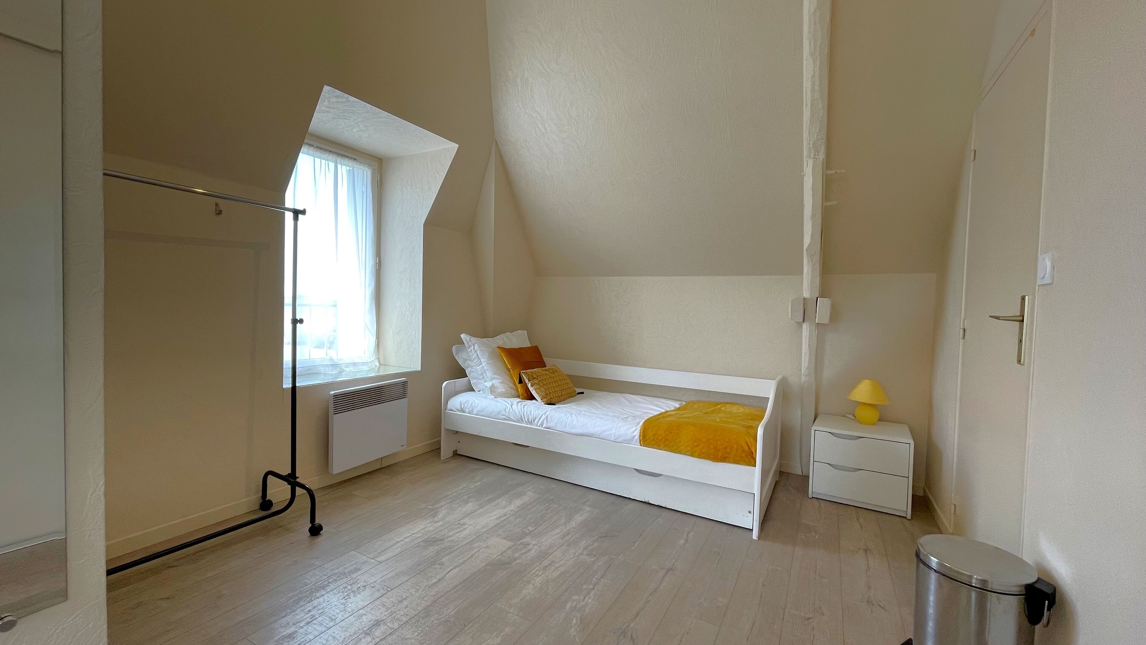 Une chambre avec un lit simple, un radiateur, une fenêtre avec des rideaux, une table de chevet avec une lampe et une poubelle.