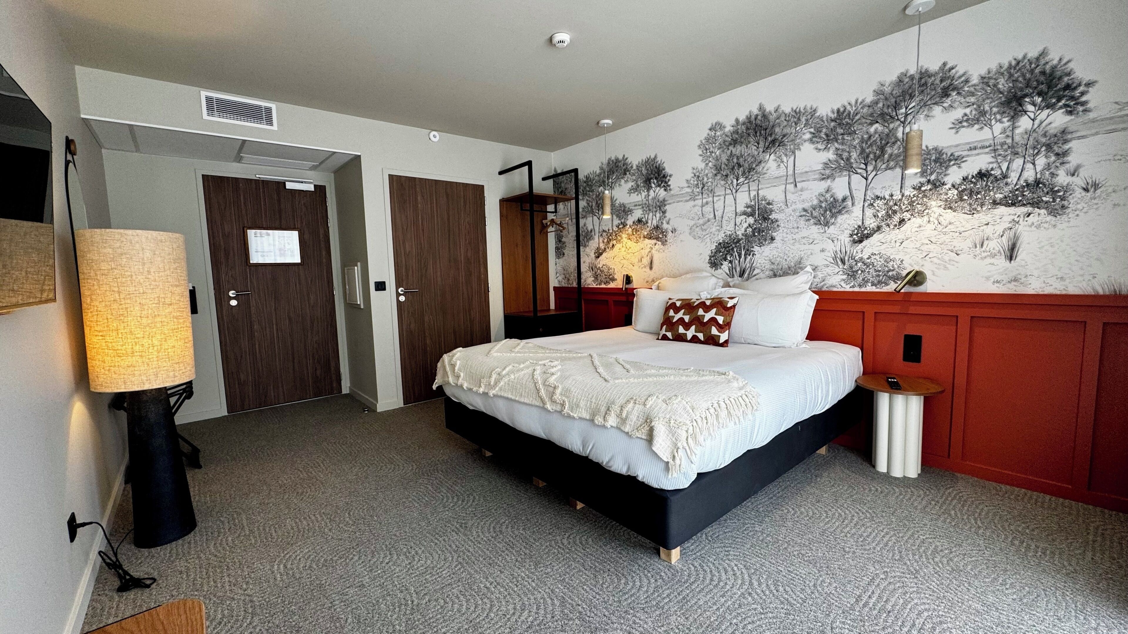 Ein Hotelzimmer mit Bett, Nachttisch, Lampe, Spiegel und Wandgemälde.