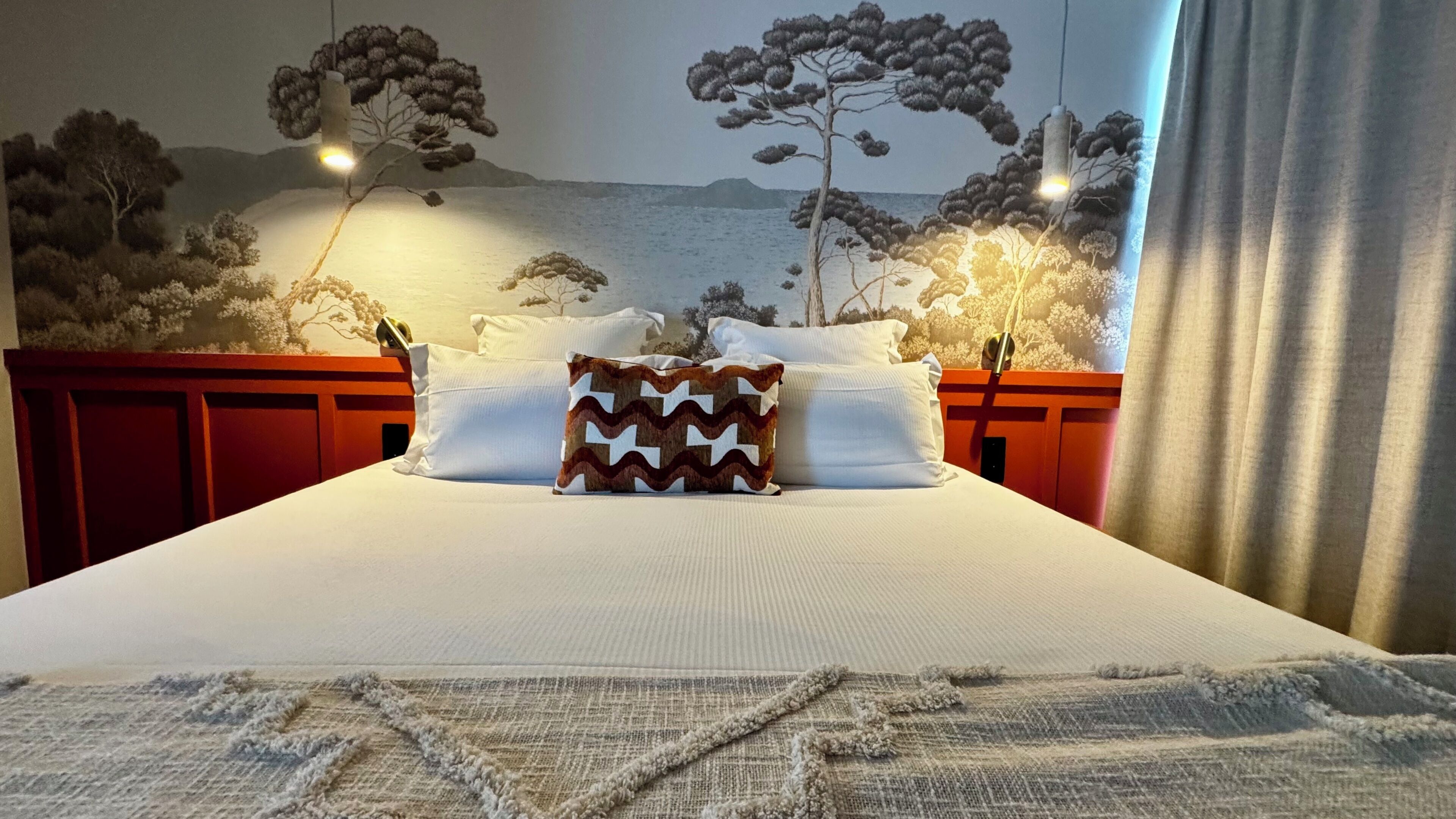 Ein Bett mit weißer Bettwäsche, einem gemusterten Kopfkissen und einer Wandmalerei mit Bäumen und einer Landschaft.