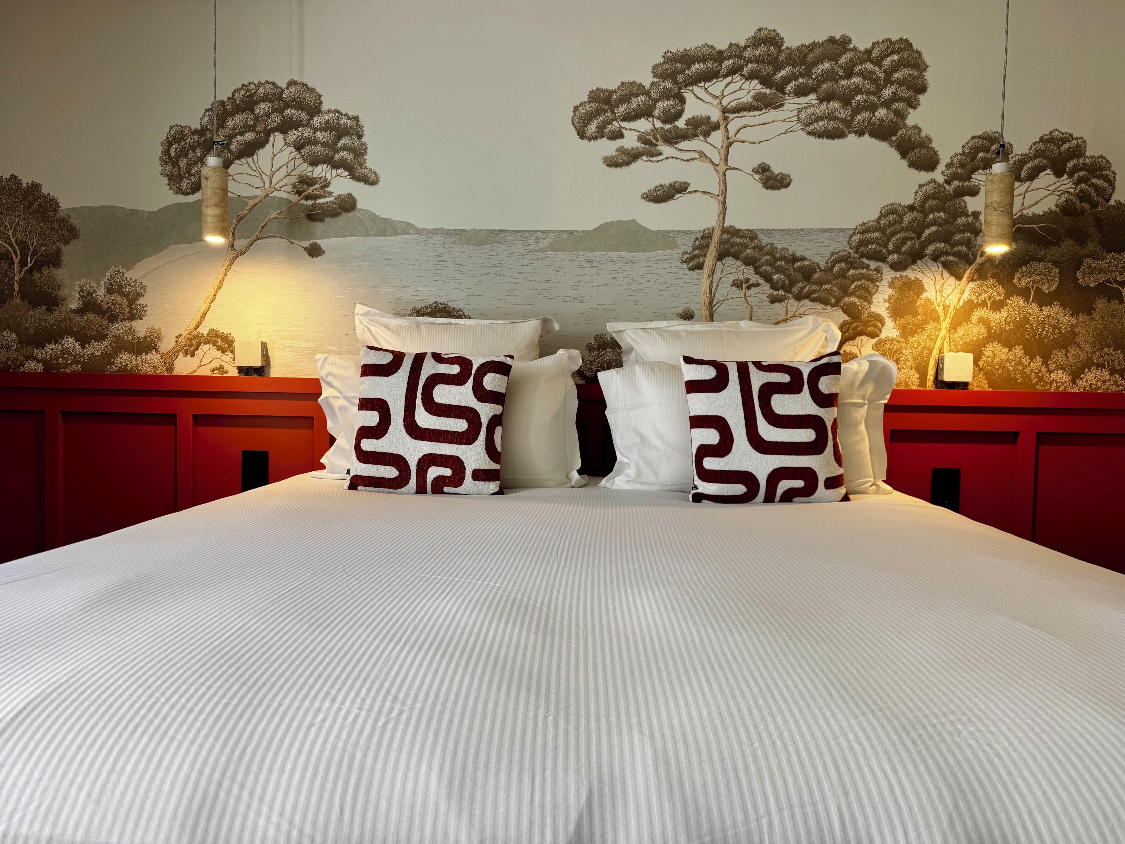 A bed with white bedding, two pillows with a geometric pattern, and a mural of trees and mountains on the wall.