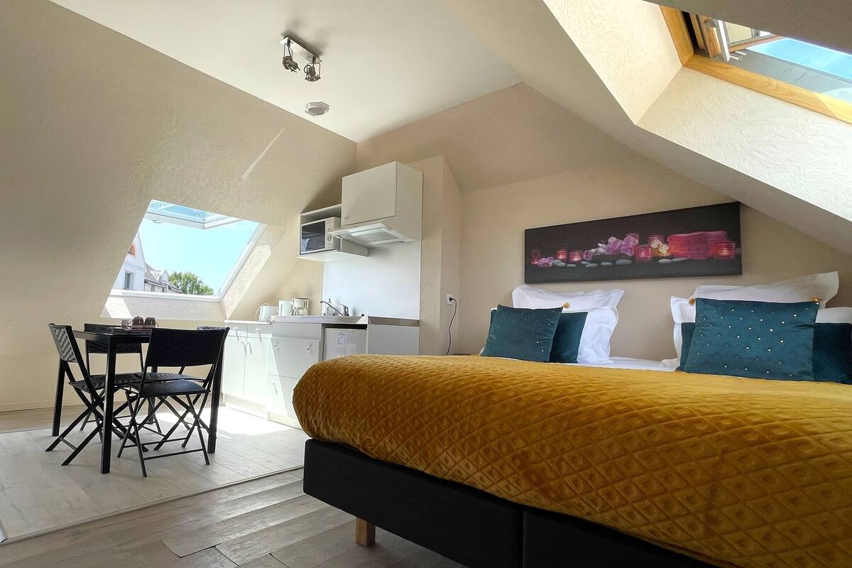 Une pièce compacte et bien éclairée, comprenant un lit, un bureau et une kitchenette.