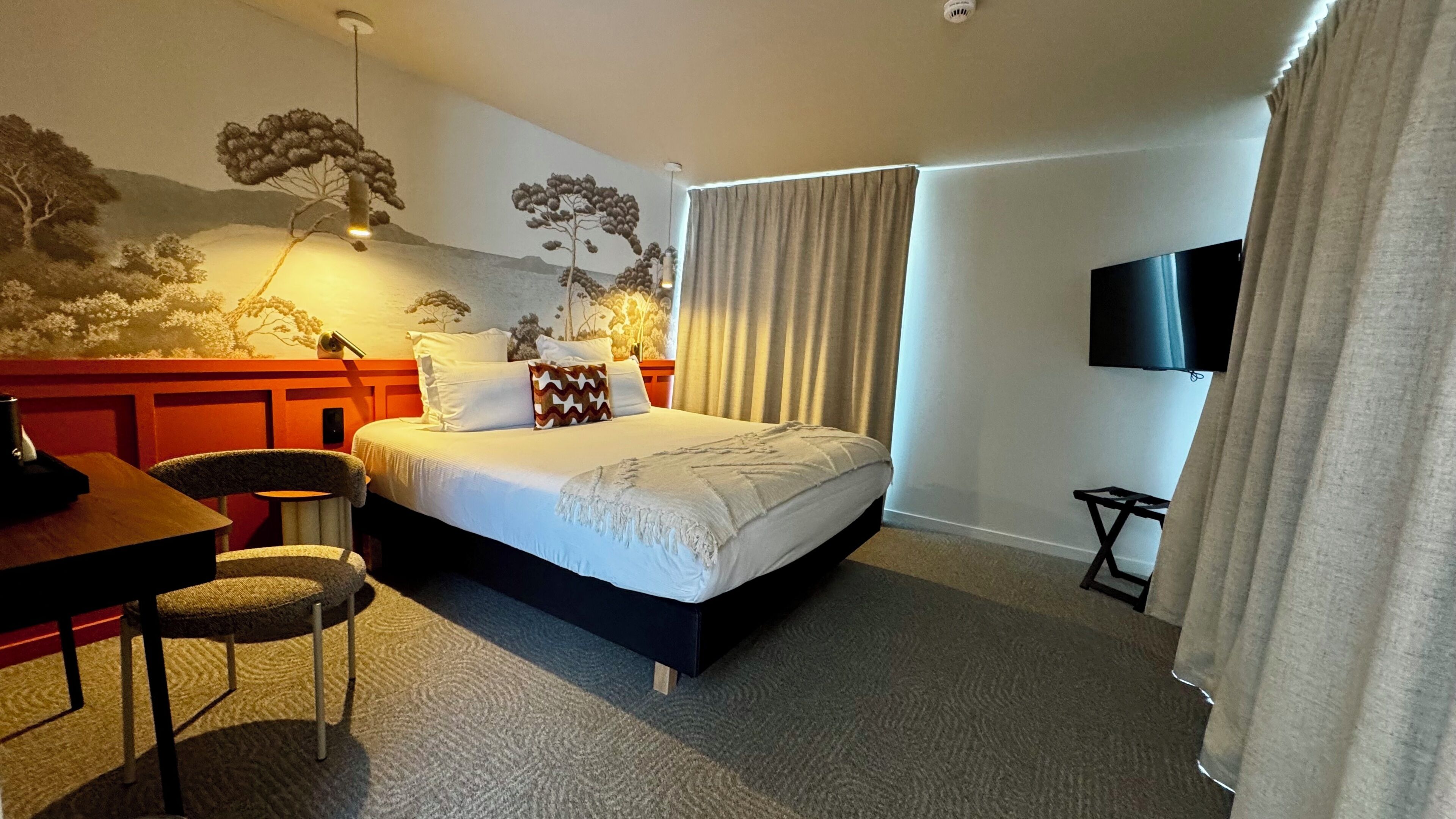 A hotel room with a bed, a desk, a chair, a television, and a wall mural.