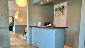 Un espace d’accueil avec un comptoir recouvert de carreaux bleu clair, un bureau d’accueil en bois et des plantes suspendues.