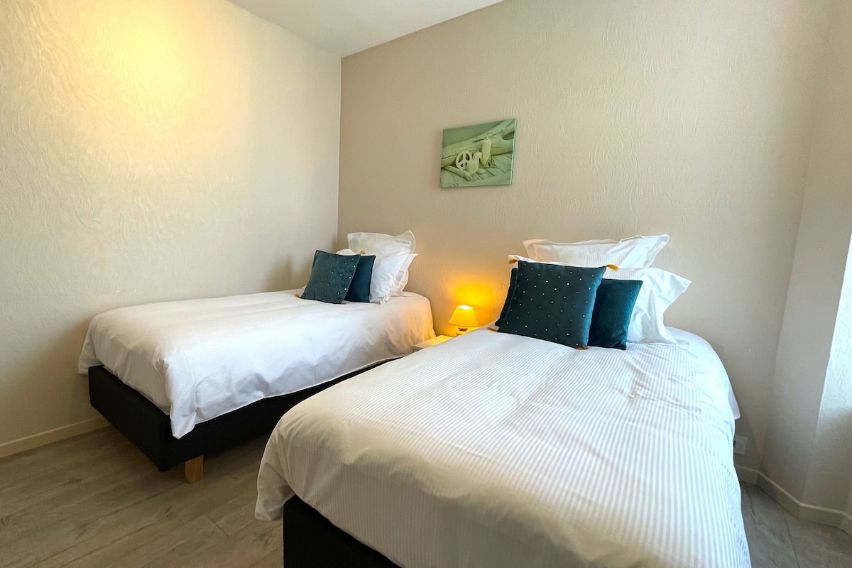 Une chambre d’hôtel avec deux lits simples, chacun étant équipé de draps blancs et d’oreillers foncés. Une petite table avec une lampe se trouve entre les deux lits.