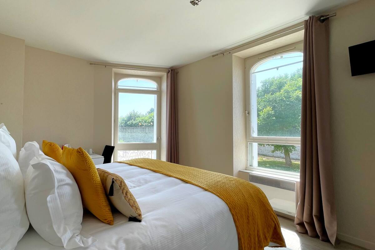 Une chambre d’hôtel avec un grand lit, deux fenêtres et une vue sur la verdure.