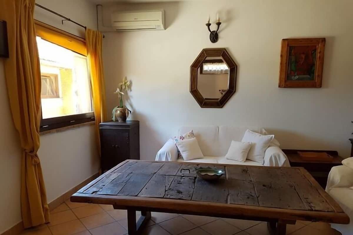 Une pièce avec une table en bois, un canapé blanc, un petit meuble, un miroir et une fenêtre avec des rideaux.