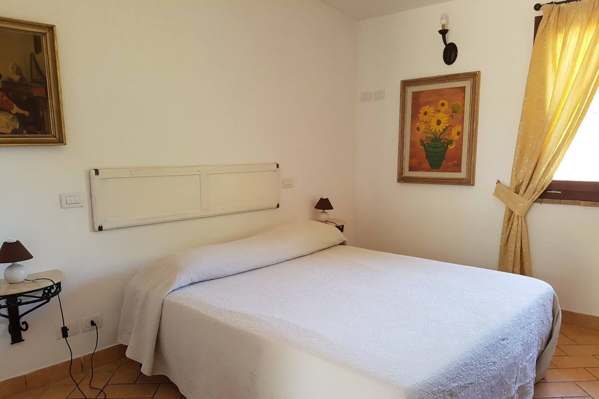 Un lit simple avec une couvre-lit blanche, une table de chevet avec une lampe, un radiateur fixé au mur et un tableau représentant des tournesols.