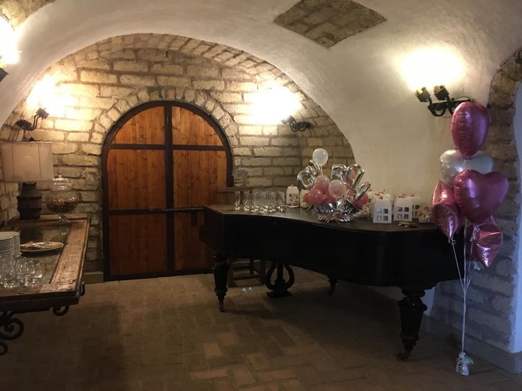 Une pièce rustique aux murs de pierre, dotée d’une porte en bois, d’une table dressée avec de la verrerie et des ballons, et d’une petite table sur laquelle sont disposées des assiettes et des verres.