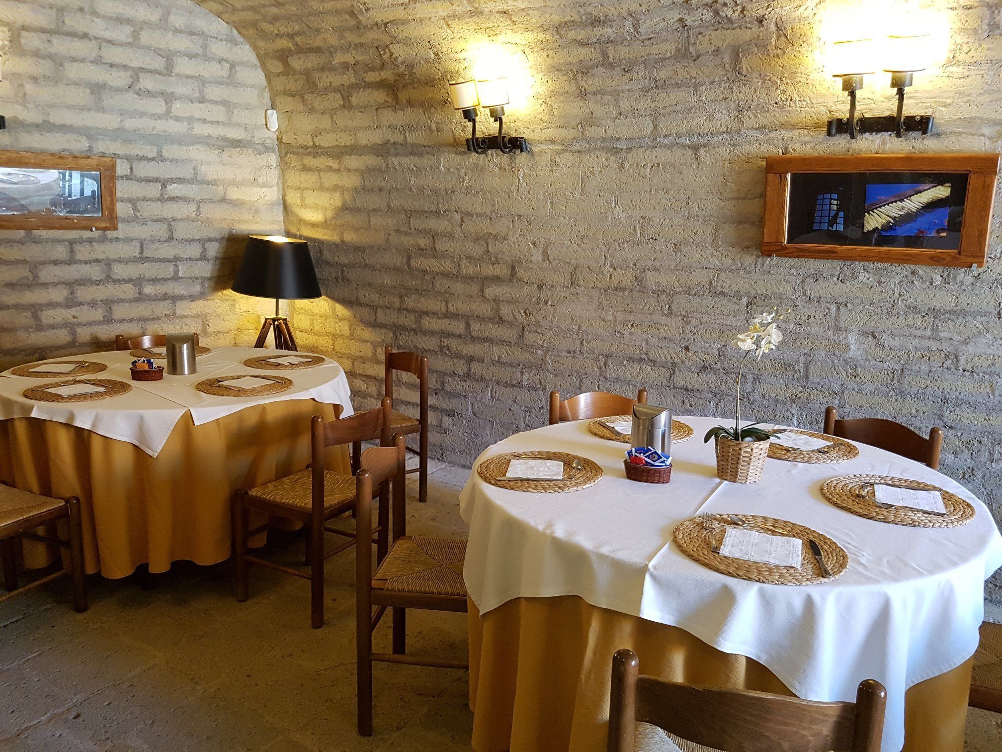 Een rustieke eetruimte met ronde tafels, bedekt met witte tafelkleden en geweven placemats, houten stoelen en een bakstenen muur.