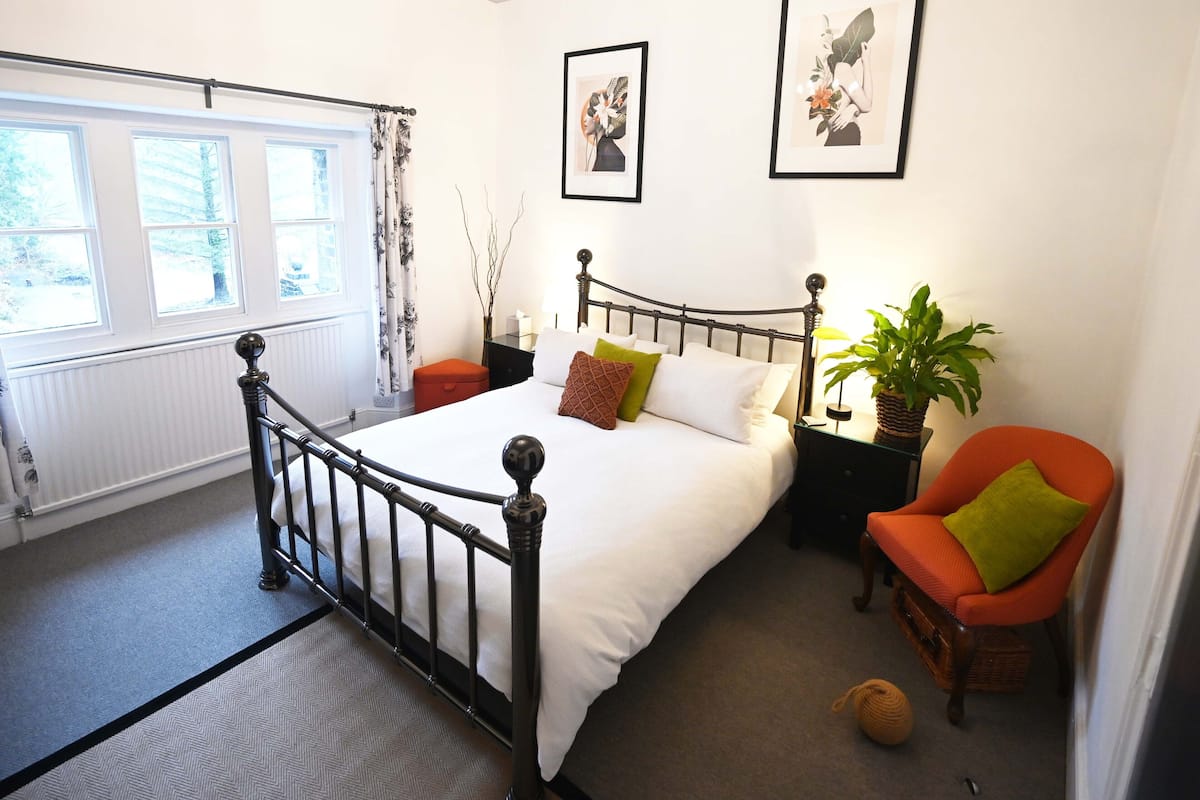 A bedroom with a bed, a nightstand, a chair, a potted plant, and two framed pictures on the wall.