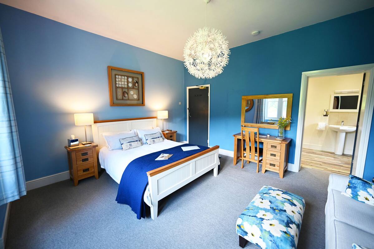 A bedroom with a bed, bedside tables, a desk, a chair, a dresser, a mirror, and a bathroom visible through an open door.