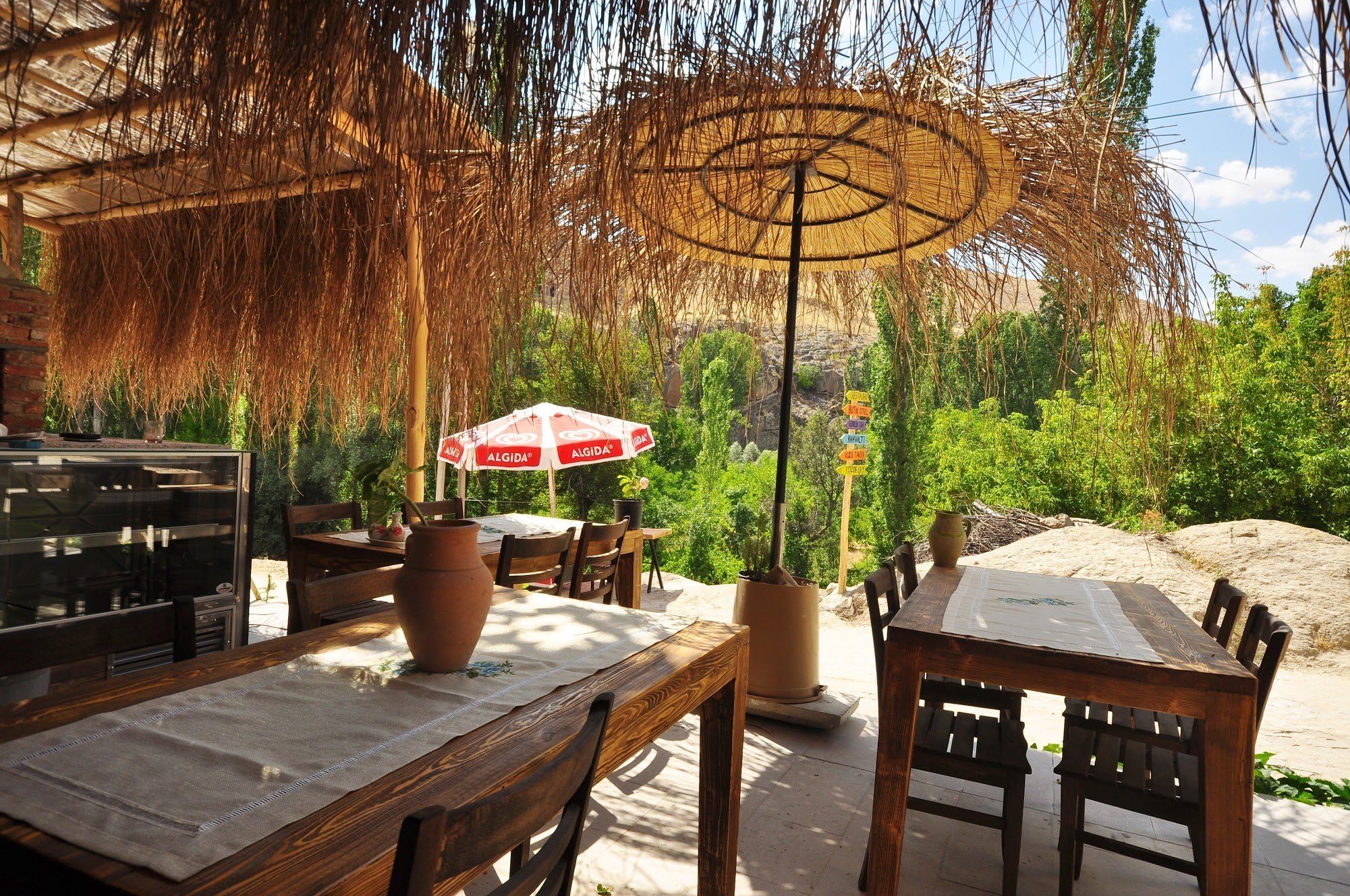 An outdoor dining area with wooden tables and chairs, a large umbrella, and a signpost with various directions.