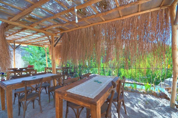 An outdoor dining area with wooden tables and chairs, a thatched roof, and a view of greenery. - BELİSIRMACAVE (Guzelyurt)