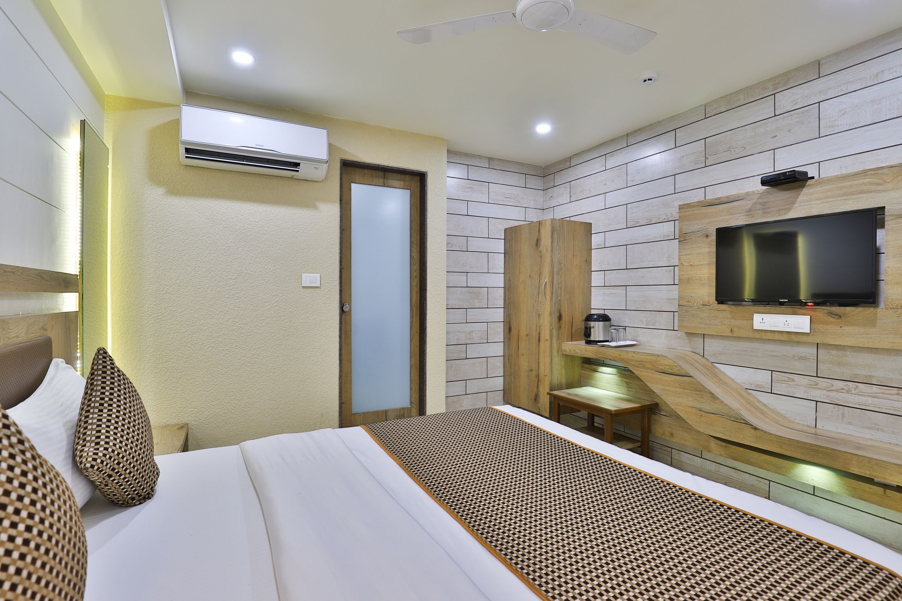 A modern hotel room with a bed, a television mounted on a wooden wall, an air conditioning unit, and a small table with a chair.