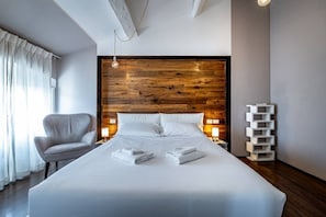 Una camera d'albergo moderna con un letto grande, una testiera in legno, una sedia bianca, lampade da comodino e un mobile contenitore bianco.