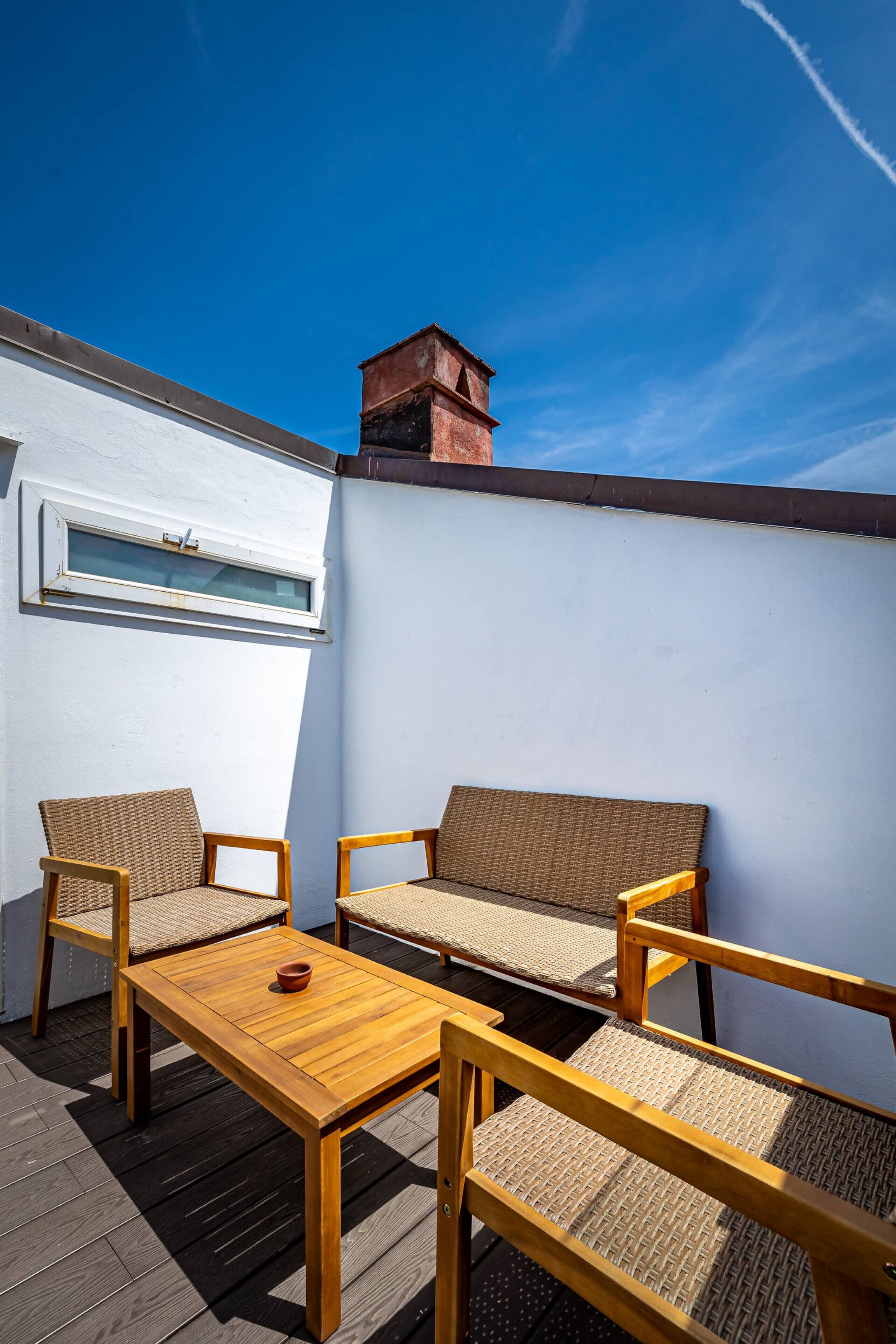 Una terraza en la azotea con dos sillas de mimbre, una mesa de madera y un ventanal.