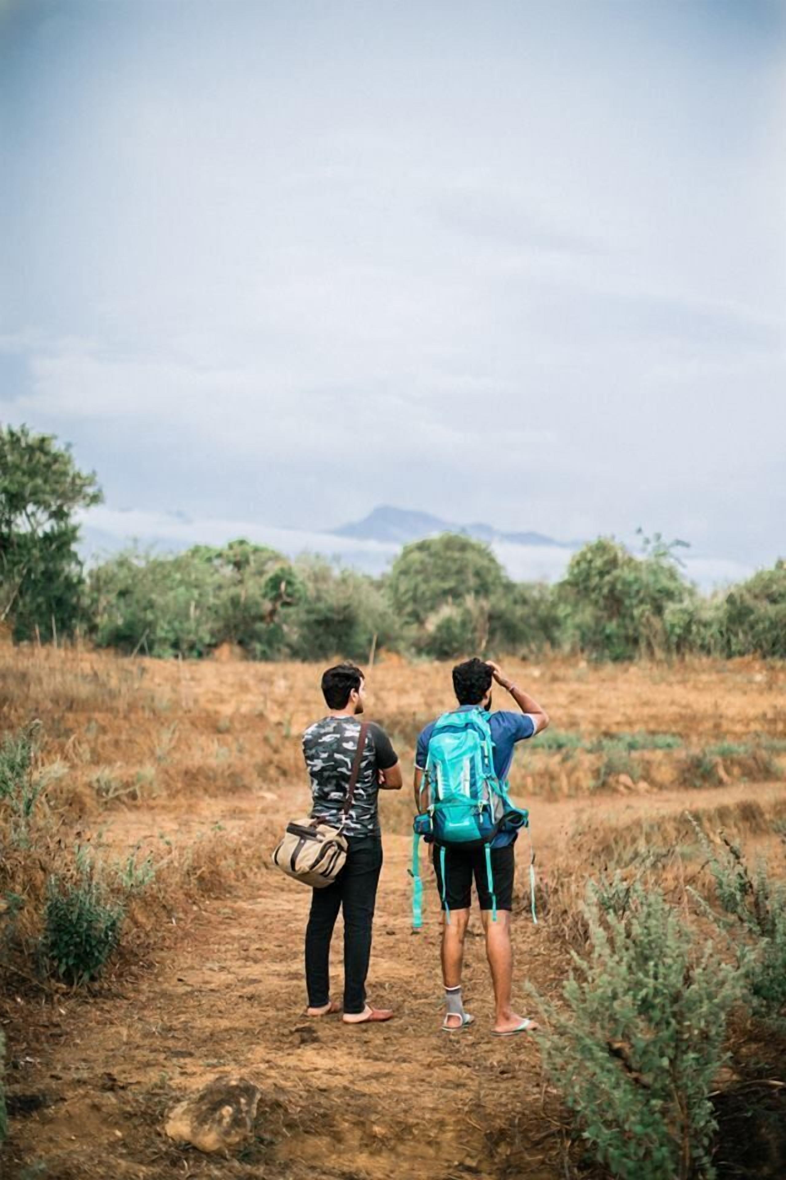 Two people standing on a dirt path in a natural setting, one holding a bag and the other with a backpack.