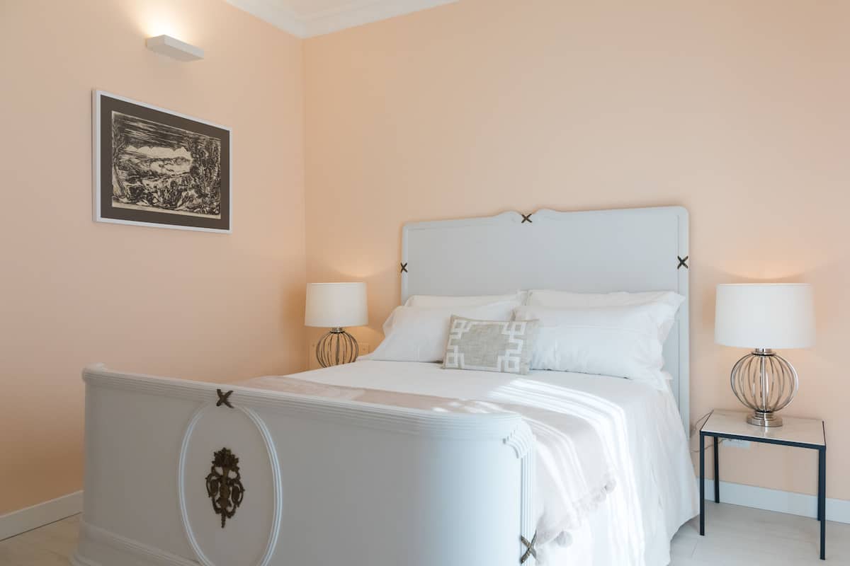 Une chambre à coucher avec un lit blanc, deux tables de chevet avec des lampes, un tableau encadré au mur et une tête de lit décorative.