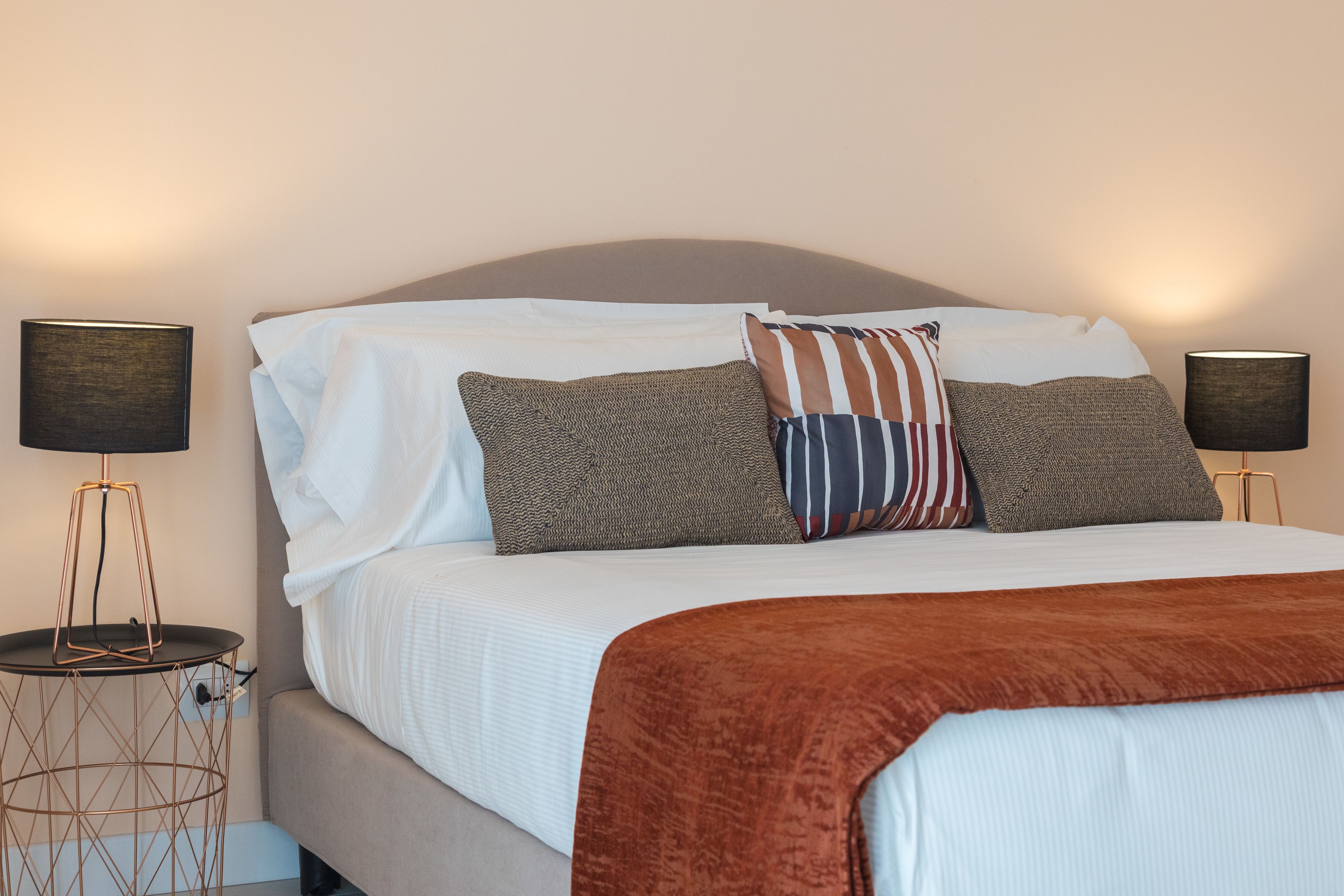 A neatly made bed with a brown throw blanket, two bedside lamps, and a variety of pillows.
