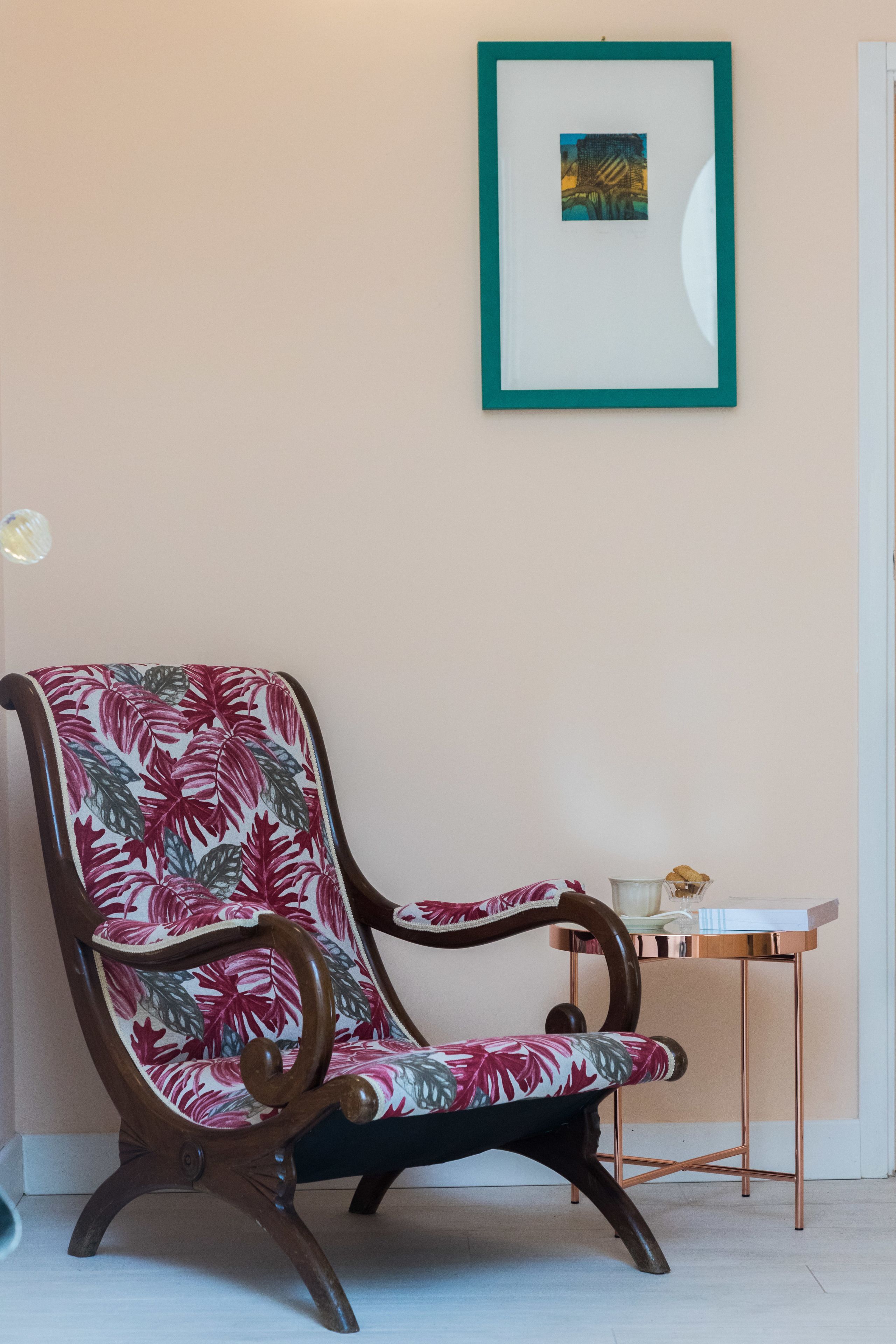 A wooden chair with a floral seat and backrest, a framed picture on the wall, and a small table with a teddy bear.
