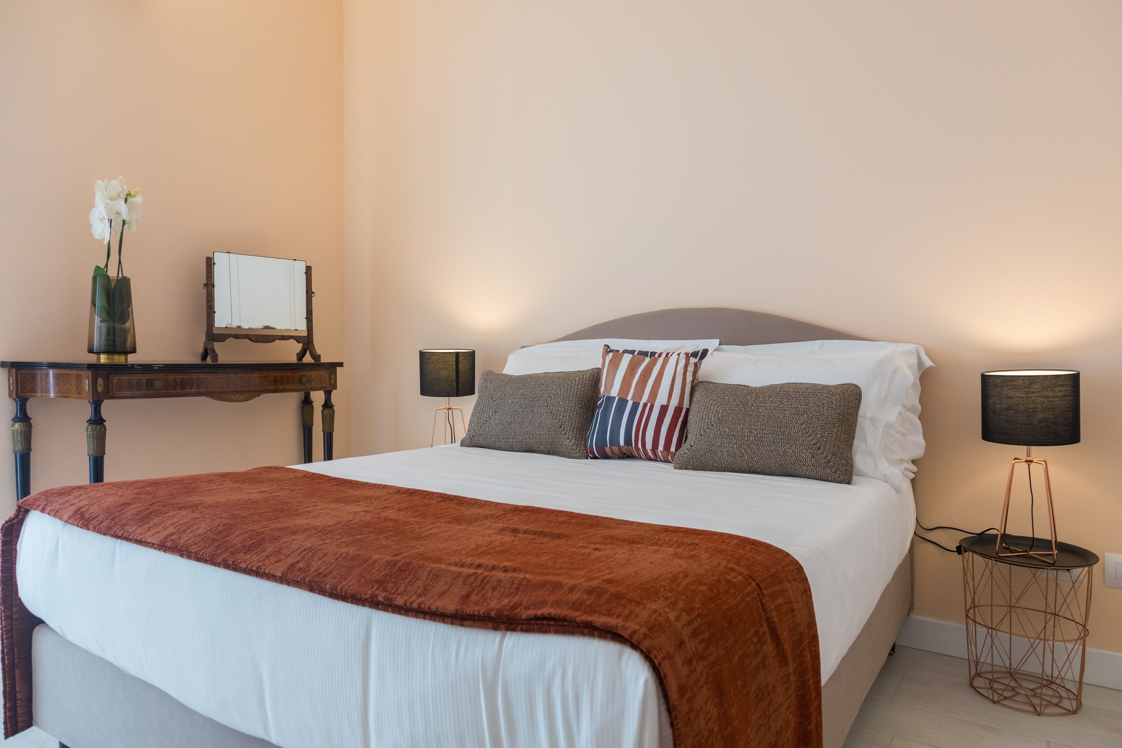 A neatly made bed with a brown blanket, two bedside lamps, a small table with a vase of flowers, and a mirror on the wall.
