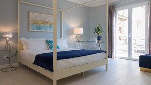 Un lit à baldaquin avec une structure blanche, un plaid bleu et une literie blanche. Une table en verre avec une lampe et une plante en pot se trouve à côté du lit. Un tableau est accroché au mur, au-dessus du lit.