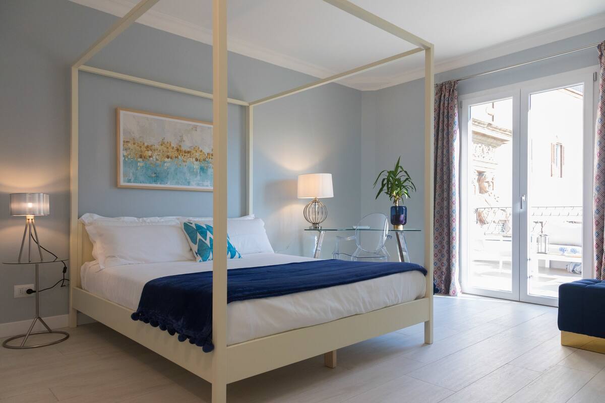 Un lit à baldaquin avec une structure blanche, un plaid bleu et une literie blanche. Une table en verre avec une lampe et une plante en pot se trouve à côté du lit. Un tableau est accroché au mur, au-dessus du lit.