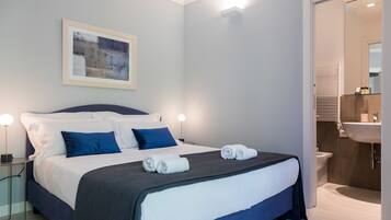 Una habitación de hotel moderna con una cama grande, una mesita de noche con lámpara, un cuadro enmarcado en la pared y un baño visible a través de una puerta abierta.
