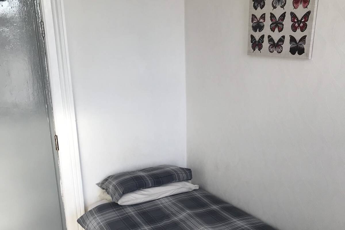 A small, single-bed room with a plaid bedspread, white curtains, and a wall-mounted butterfly decoration.