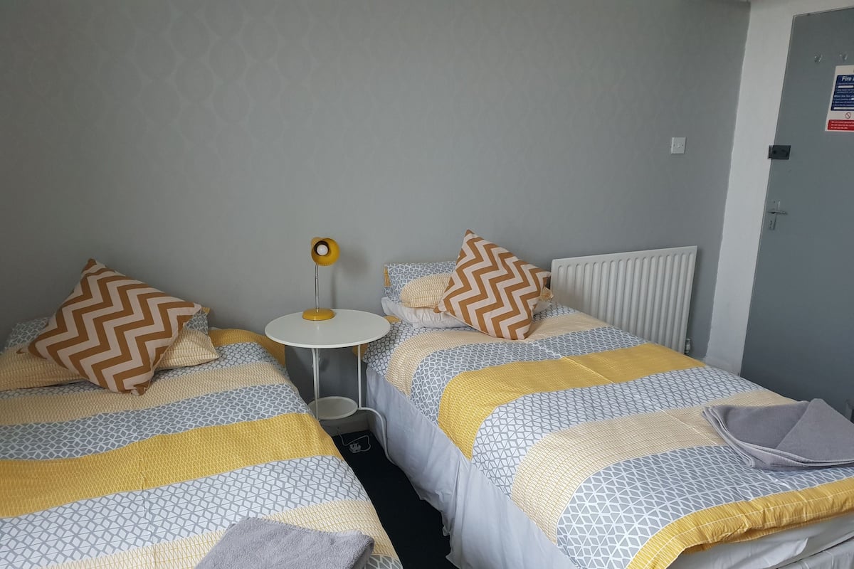 A hotel room with two beds, patterned bedding, a small round table with a lamp, and a radiator.