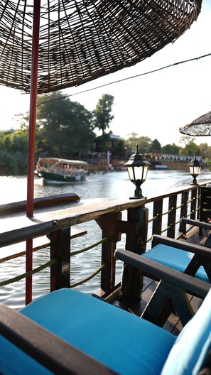 A wooden deck with blue cushioned seating, a woven canopy overhead, and a view of a river with boats and buildings in the background.