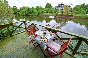 A dining table set on a wooden deck with a view of a calm lake and a house in the background.