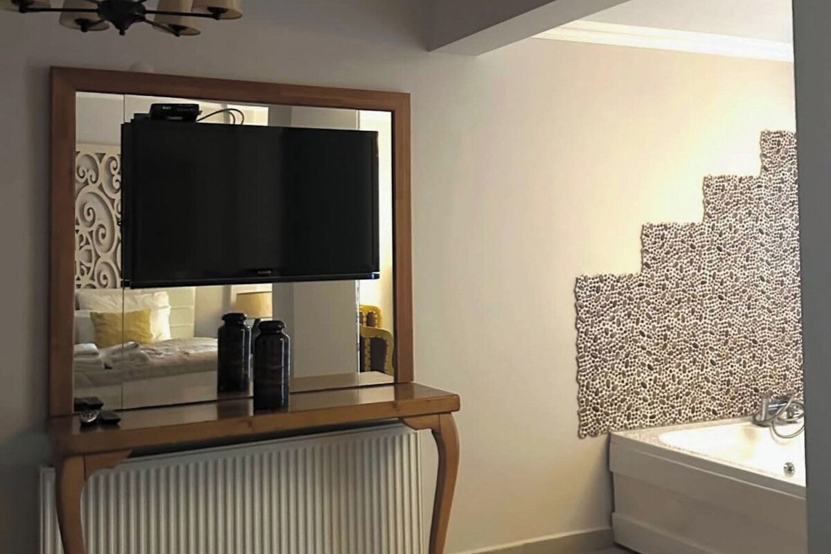 A hotel room with a large mirror, flat-screen TV, radiator, and a bathroom with a sink and towel stack.