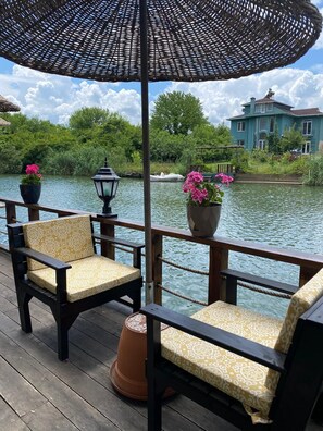 A wooden deck with two chairs and a large umbrella, overlooking a body of water with a house in the background.