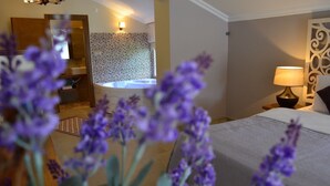 A hotel room with a bed, a nightstand with a lamp, a bathroom with a shower and sink, and purple flowers in the foreground. - Sweet Home River Hotel (Sile)