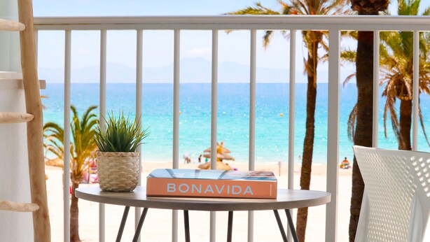 A balcony with a view of the beach, a book titled 'BONAVIDA', and a potted plant.