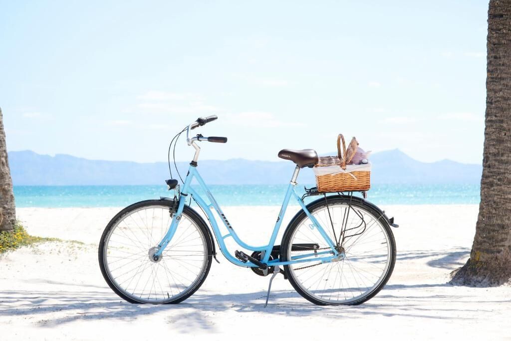 Una bicicleta azul claro con una cesta de mimbre en una playa arenosa con palmeras y un cielo despejado.