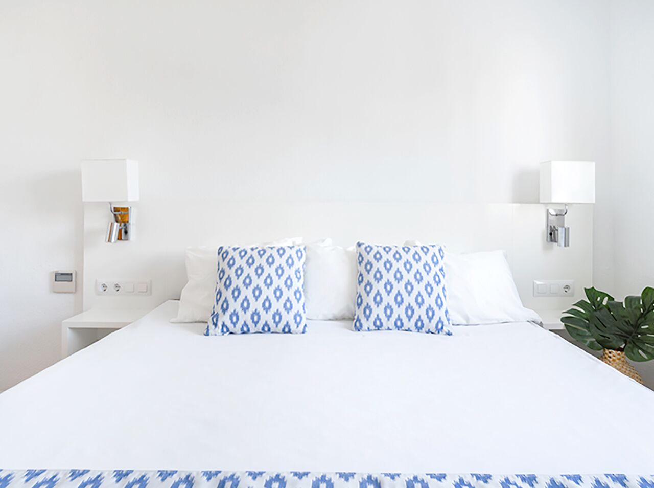A hotel room with a large bed, white bedding, two patterned pillows, bedside lamps, a small table, and a potted plant.